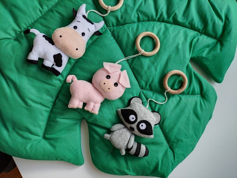 May include: A green felt play mat with a cow, pig, and raccoon hanging from wooden rings. The animals are made of felt and have embroidered details.