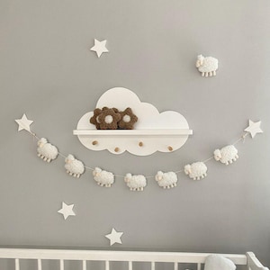 May include: A nursery wall decor set featuring a white cloud-shaped shelf with two brown flower-shaped decorations. A garland of white sheep hangs below, with star-shaped wall decorations. The decor is set against a gray wall.
