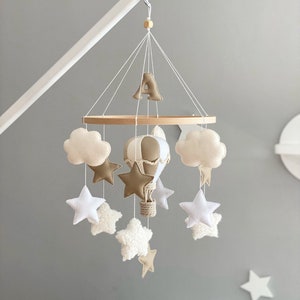 Puede incluir: Un móvil de bebé blanco y beige con un globo aerostático, nubes y estrellas. El móvil está colgado de un brazo blanco y tiene la letra 'A' en la parte superior.