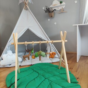 May include: A wooden play gym with hanging felt animal toys, a green leaf-shaped play mat, and a gray teepee tent. The play gym is set up in a room with a wooden shelf with a cloud design and a basket of dried flowers.