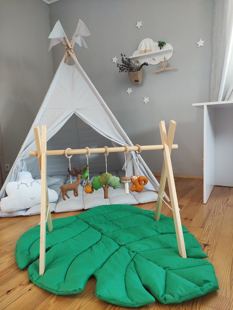May include: A wooden play gym with hanging felt animal toys, a green leaf-shaped play mat, and a gray teepee tent. The play gym is set up in a room with a wooden shelf with a cloud design and a basket of dried flowers.