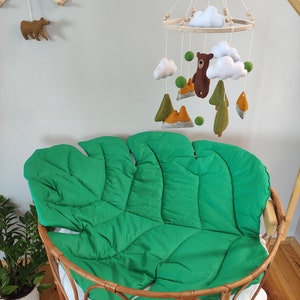 May include: A green felt leaf-shaped baby nest with a wooden frame. A wooden mobile with felt clouds, mountains, trees, and a bear hangs above the nest.