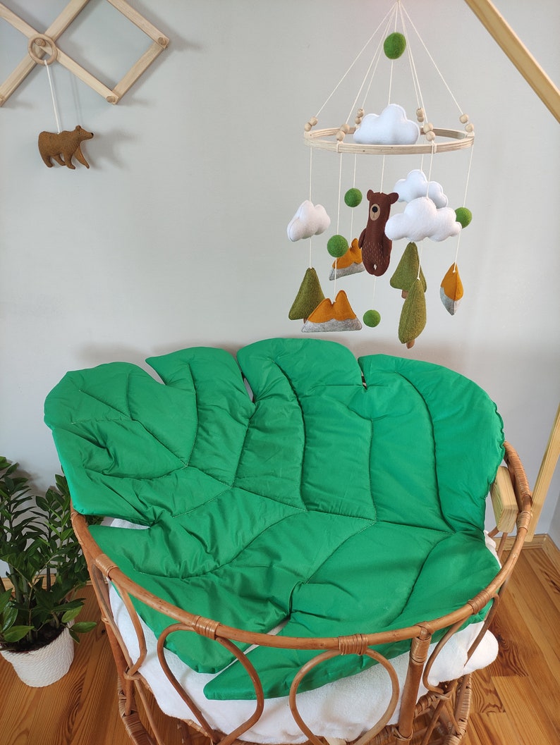 May include: A green felt leaf-shaped baby nest with a wooden frame. A wooden mobile with felt clouds, mountains, trees, and a bear hangs above the nest.