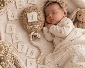 May include: A beige crocheted balloon with the name Arian embroidered on it, accompanied by twelve square felt cards numbered one through twelve. The balloon features a rope string and a square felt card with the number one attached.
