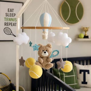 May include: A nursery mobile featuring a central light blue and white hot air balloon, a brown teddy bear holding a glittery blue tennis racket, yellow tennis balls, white clouds, and glittery blue and grey stars suspended from a wooden ring.
