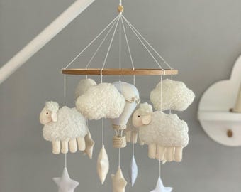 May include: A baby mobile featuring fluffy white sheep, clouds, stars, and a hot air balloon. The mobile is suspended from a wooden ring and hangs from a white arm. The nursery decor is in soft neutral colors.