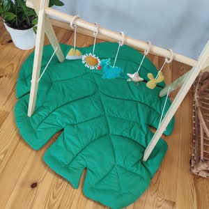 May include: A wooden play gym with a green monstera leaf play mat and hanging toys. The toys include a yellow sun, a blue and green dragon, a gold star, and a yellow and green cactus.