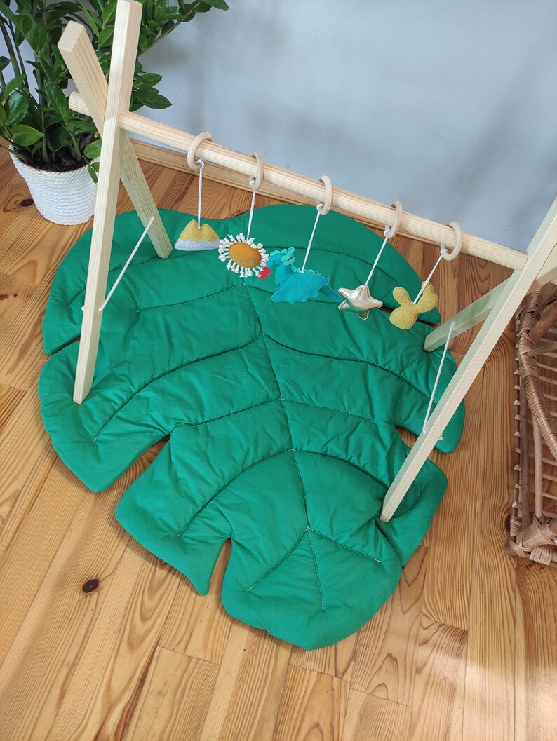 May include: A wooden play gym with a green monstera leaf play mat and hanging toys. The toys include a yellow sun, a blue and green dragon, a gold star, and a yellow and green cactus.