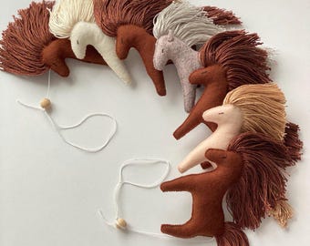 May include: A collection of felt horse ornaments in various colors, including cream, brown, and pink, with long, fringed manes and tails. Each horse is suspended by a white string with a wooden bead, creating a whimsical mobile or decorative piece.
