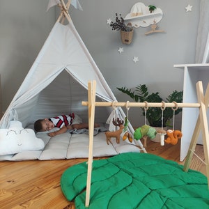May include: A white teepee tent with a gray and white quilted mattress inside. A child is lying down inside the tent. A wooden play gym with hanging toys is in the foreground. The toys include a deer, a mushroom, a squirrel, and a tree. A green leaf-shaped play mat is on the floor.