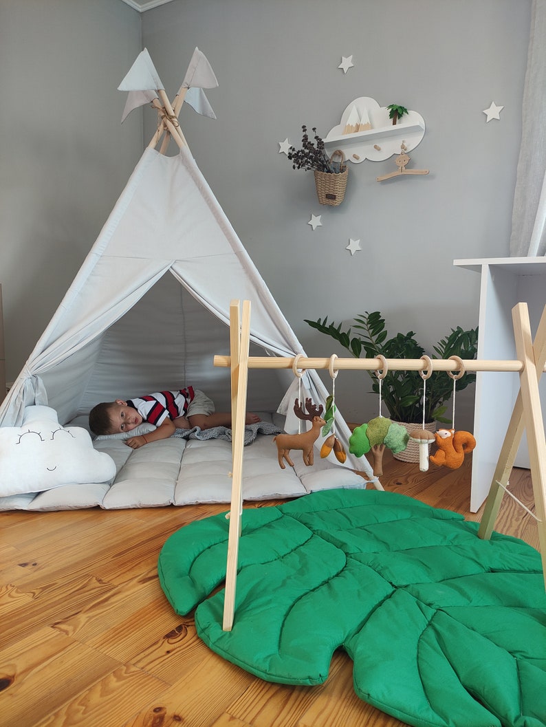 May include: A white teepee tent with a gray and white quilted mattress inside. A child is lying down inside the tent. A wooden play gym with hanging toys is in the foreground. The toys include a deer, a mushroom, a squirrel, and a tree. A green leaf-shaped play mat is on the floor.