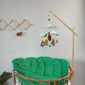 May include: A wooden mobile arm with a felt woodland animal mobile hanging from it. The mobile features a bear, fox, and trees. The mobile is hanging over a green crib mattress.