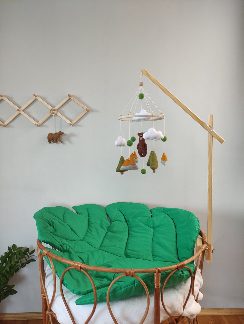 May include: A wooden mobile arm with a felt woodland animal mobile hanging from it. The mobile features a bear, fox, and trees. The mobile is hanging over a green crib mattress.