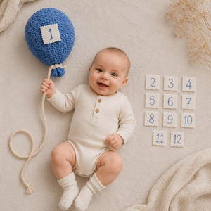 May include: A baby wearing a cream-coloured long-sleeve ribbed bodysuit and matching socks. The baby holds a blue textured balloon with a white square patch featuring the number 1. Eleven additional white square patches numbered 2 through 12 are nearby.