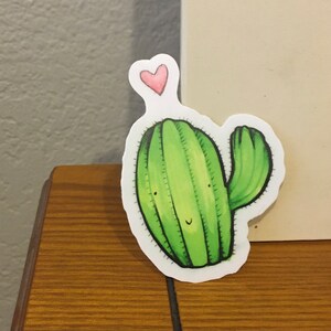 May include: A green cactus with a pink heart on top. The cactus has a smiling face and a single arm.