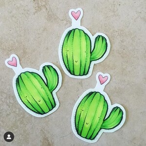 May include: Three green cactus stickers with pink hearts on top. Each cactus has a smiling face and a single arm.