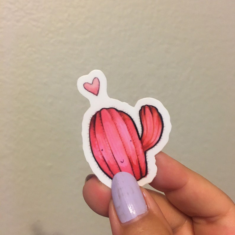Pode incluir: Um adesivo de cacto rosa com um pequeno cora&ccedil;&atilde;o rosa acima. O cacto tem um rosto sorridente e um pequeno bra&ccedil;o.