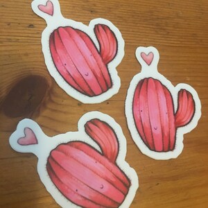 Pode incluir: Tr&ecirc;s adesivos de cacto rosa com um cora&ccedil;&atilde;o acima de cada um. Os cactos s&atilde;o desenhados em estilo de desenho animado com um rosto sorridente.