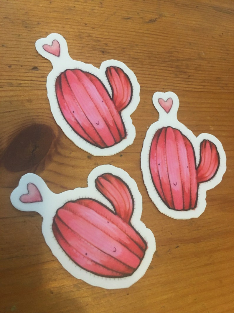 Pode incluir: Tr&ecirc;s adesivos de cacto rosa com um cora&ccedil;&atilde;o acima de cada um. Os cactos s&atilde;o desenhados em estilo de desenho animado com um rosto sorridente.
