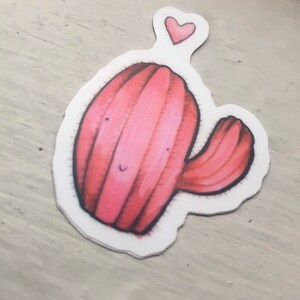 Pode incluir: Um cacto rosa com um rosto sorridente e um pequeno cora&ccedil;&atilde;o acima dele. O cacto tem um &uacute;nico bra&ccedil;o que se estende para o lado.