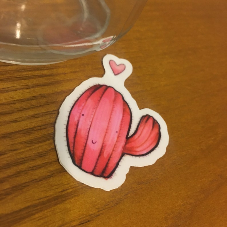 Pode incluir: Um adesivo de cacto rosa com um pequeno cora&ccedil;&atilde;o acima dele. O cacto tem um rosto sorridente e um &uacute;nico bra&ccedil;o.