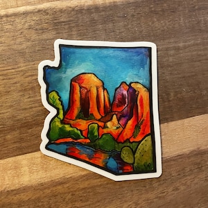 May include: A colorful sticker featuring a stylized landscape with red rock formations, green trees, and a blue body of water. The sticker is shaped like the state of Arizona.