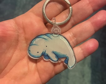 May include: A light blue manatee keychain with a silver key ring. The manatee is made of clear acrylic with a hand-painted design.