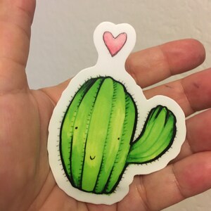May include: A green cactus sticker with a smiling face and a pink heart on top. The sticker is on a white background.