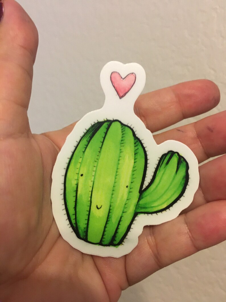 May include: A green cactus sticker with a smiling face and a pink heart on top. The sticker is on a white background.