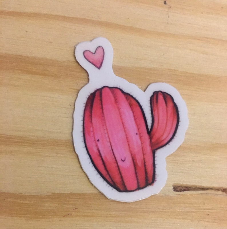 Pode incluir: Um adesivo de cacto rosa com um pequeno cora&ccedil;&atilde;o rosa no topo. O cacto tem um rosto sorridente e um &uacute;nico bra&ccedil;o.