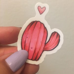 Pode incluir: Um adesivo de cacto rosa com um pequeno cora&ccedil;&atilde;o rosa acima. O cacto tem um rosto sorridente e um &uacute;nico bra&ccedil;o.