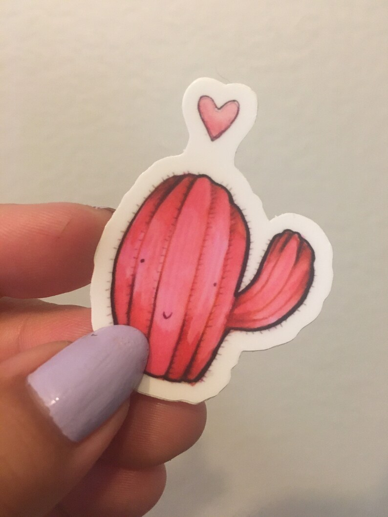 Pode incluir: Um adesivo de cacto rosa com um pequeno cora&ccedil;&atilde;o rosa acima. O cacto tem um rosto sorridente e um &uacute;nico bra&ccedil;o.