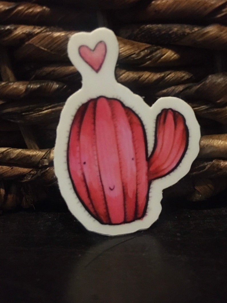 Pode incluir: Um adesivo de cacto rosa com um pequeno cora&ccedil;&atilde;o no topo. O cacto tem um rosto sorridente e um &uacute;nico bra&ccedil;o.