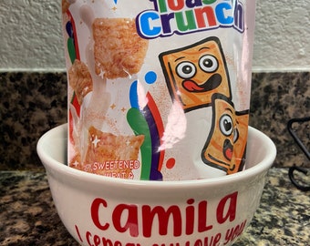 May include: A white ceramic bowl with the words "Camila Cereal-sly Love You" in red lettering. A bag of Cinnamon Toast Crunch cereal is sitting inside the bowl.