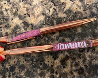 Puede incluir: Dos bolígrafos de purpurina de color oro rosa con barriles transparentes. Un bolígrafo tiene el nombre "Tamara" escrito en letras blancas.