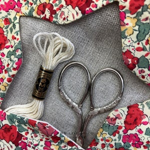 May include: A white embroidery floss spool with a gold label that reads "DMC 25 8m (8.7 yds)" and a pair of silver embroidery scissors on a gray fabric background with a floral pattern.