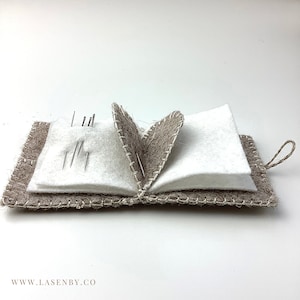 Op de afbeelding: Open vilten naaldenboek met naalden. Het boek is gemaakt van bruinachtig vilt met witte vilten pagina's en witte stiksels. Het naaldenboek heeft een lus om op te hangen. De website www.lasenby.co is zichtbaar.