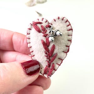 Pode incluir: Um enfeite de feltro em forma de coração com costuras e bordados vermelhos. Um pingente de bolota prateada pende do topo. O enfeite é branco-sujo com detalhes bordados em vermelho e uma borda costurada em vermelho.