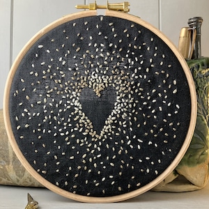 May include: A black embroidery hoop featuring a white and gold heart design. The heart is created using small stitches, giving it a textured appearance.