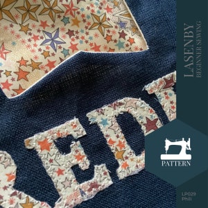 May include: A navy blue fabric with the word "READY" spelled out in white fabric with a star pattern. The fabric is stitched together with white thread. The image also includes a sewing machine icon and the text "LASENBY BEGINNER SEWING PATTERN LP029 Phili".