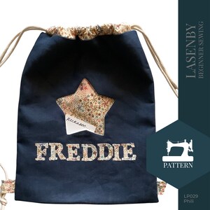 May include: A navy blue drawstring bag with a star-shaped window revealing a colorful fabric with stars. The bag has the name "FREDDIE" embroidered on the front and the text "peekaboo..." inside the star.