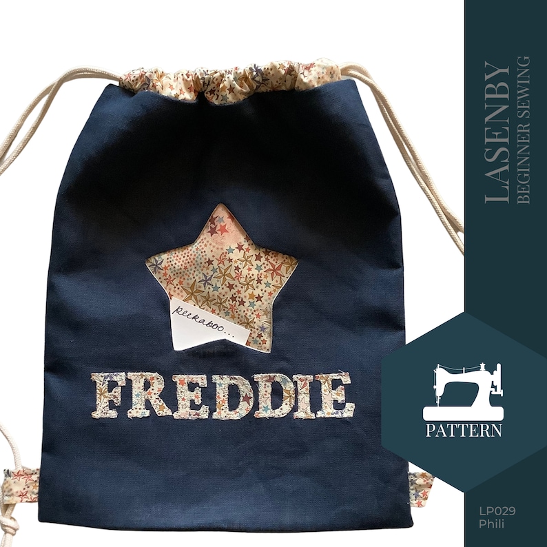 May include: A navy blue drawstring bag with a star-shaped window revealing a colorful fabric with stars. The bag has the name "FREDDIE" embroidered on the front and the text "peekaboo..." inside the star.