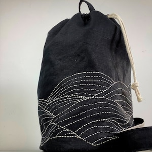 Pode incluir: Mochila preta com cordão e um padrão de ondas brancas bordado na parte inferior. A bolsa tem um fecho de cordão de cor natural e uma alça para transportar. Feita de tecido texturizado.