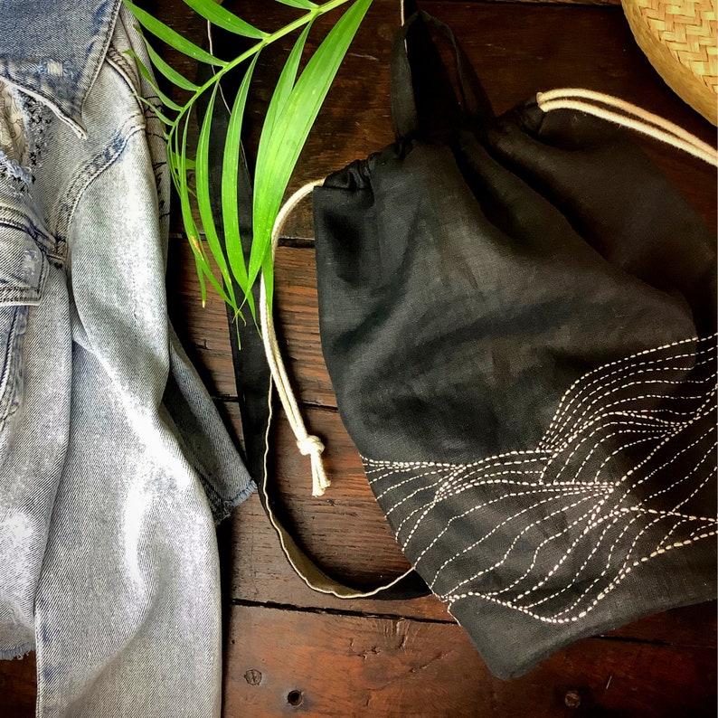 May include: A black drawstring backpack with white embroidered wave-like design. The bag has a cream-colored drawstring and strap. A light blue denim jacket and green leaf are also in the image, set against a dark wood background.