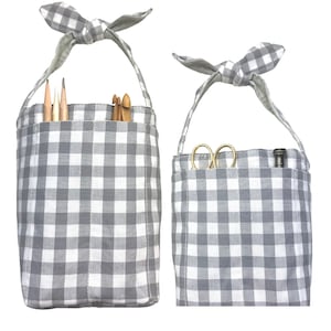 May include: Two gray and white gingham fabric bags with handles. The larger bag has a pocket with pencils and knitting needles inside. The smaller bag has a pocket with scissors and a tool inside.