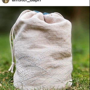 May include: A beige linen drawstring bag with a decorative stitched pattern. The bag has a light blue interior and a matching drawstring closure. The bag is sitting on a bed of green grass.