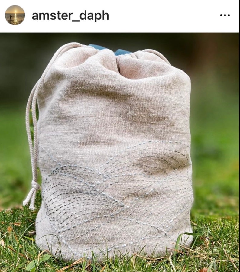 May include: A beige linen drawstring bag with a decorative stitched pattern. The bag has a light blue interior and a matching drawstring closure. The bag is sitting on a bed of green grass.