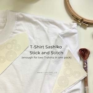 以下が含まれることがあります： ステッチの円が描かれた白い紙2枚、白いTシャツ、木製刺繍枠、茶色の刺繍糸の糸巻き。紙には「T-Shirt Sashiko Stick and Stitch (enough for two T-shirts in one pack)」と「WWW.LASENBY.CO LONDON」と書かれています。