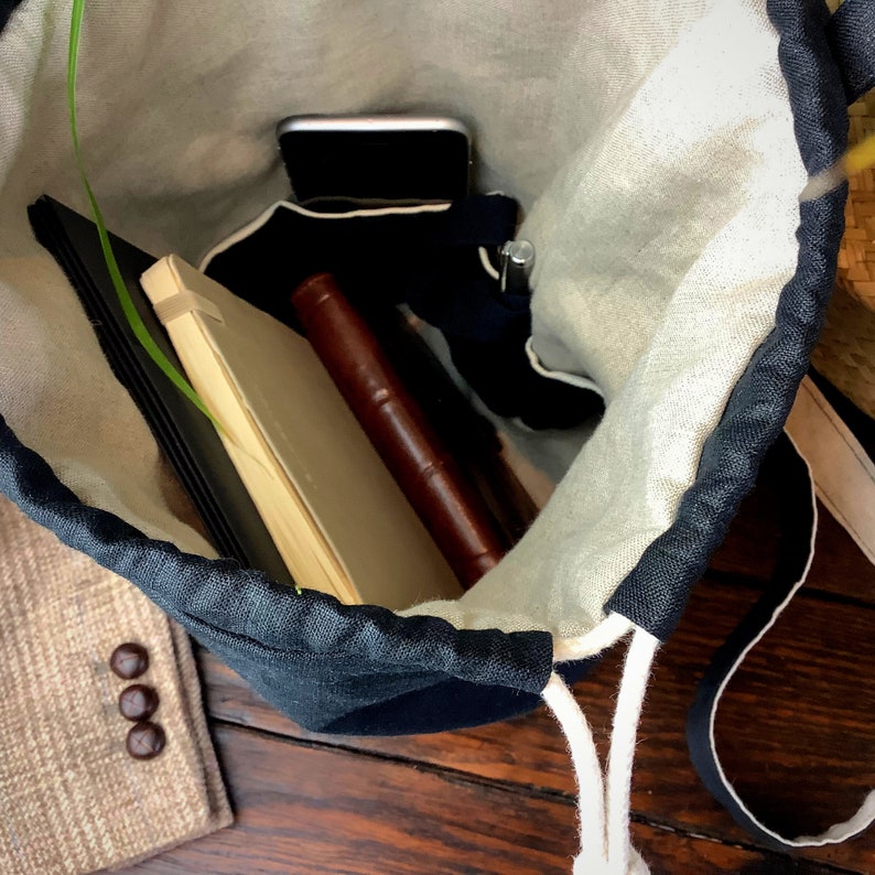 May include: A navy blue and cream drawstring bag filled with a smartphone, notebooks, and a pen. The bag is made of linen and sits on a wooden surface. A tan blazer with three buttons is visible in the corner.