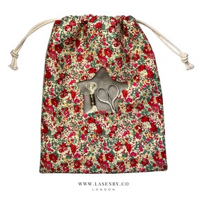 May include: A white drawstring bag with a red and green floral print. The bag has a star-shaped opening with a white thread and scissors inside. The bag has the text "www.lasenby.co LONDON" printed on the bottom.